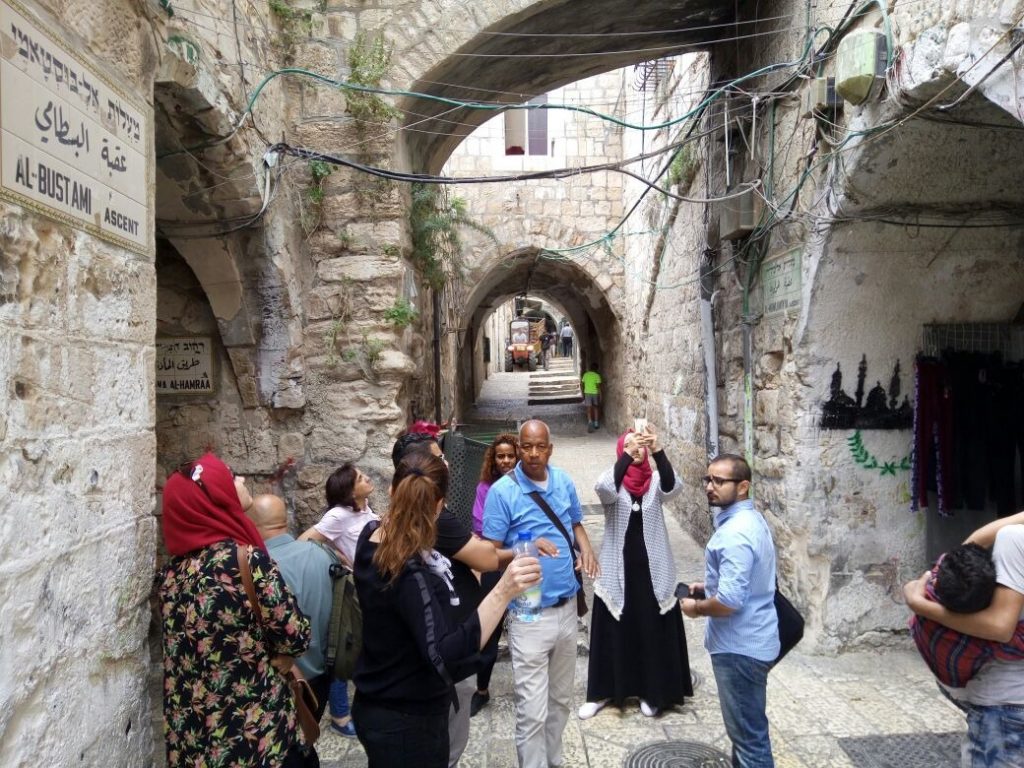 A photograph of Mahmoud Jaddeh, a middle-aged Black man wearing a blue shirt, light coloured trousers and a satchel, guiding a group of about ten tourists through the arched alleyways of the African Quarter in Jerusalem.