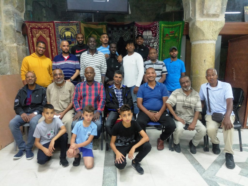 A group photograph of men and boys of varying ages from the Afro-Palestinian community, lined up in rows and smiling or just looking at the camera. They are inside a stone building with patterned fabrics hanging on the wall behind them.