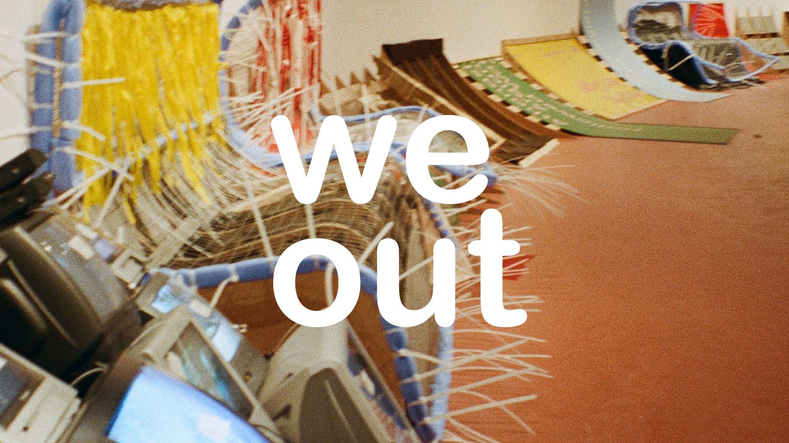 The text 'we out' sits on top of a film image taken of RESOLVE's exhibition 'them's the breaks'. There is a series of makeshift chairs and installation furniture lined up against a curved wall.
