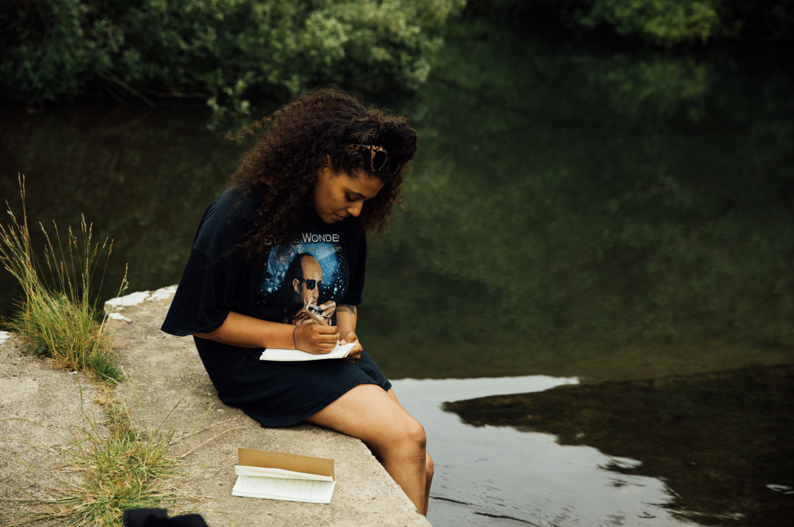 A photograph showing Evie Muir seated on the edge of a river or lake, her feet dangling in the dark water as she is bent over a notebook, pen in hand. She is wearing a t-shirt with Stevie Wonder on it and has sunglasses pushed up onto her head.